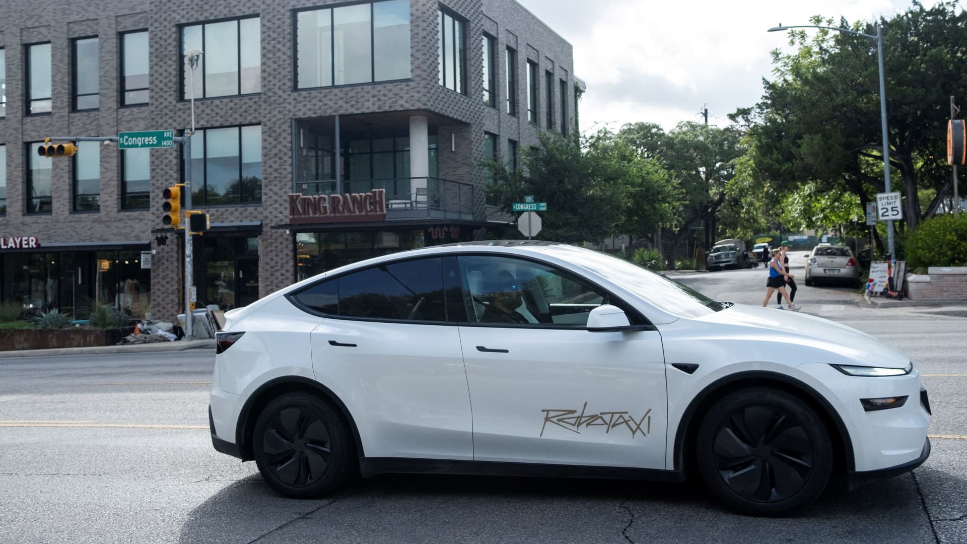 A white Tesla Model Y robotaxi with no driver at the wheel navigating a downtown Austin street